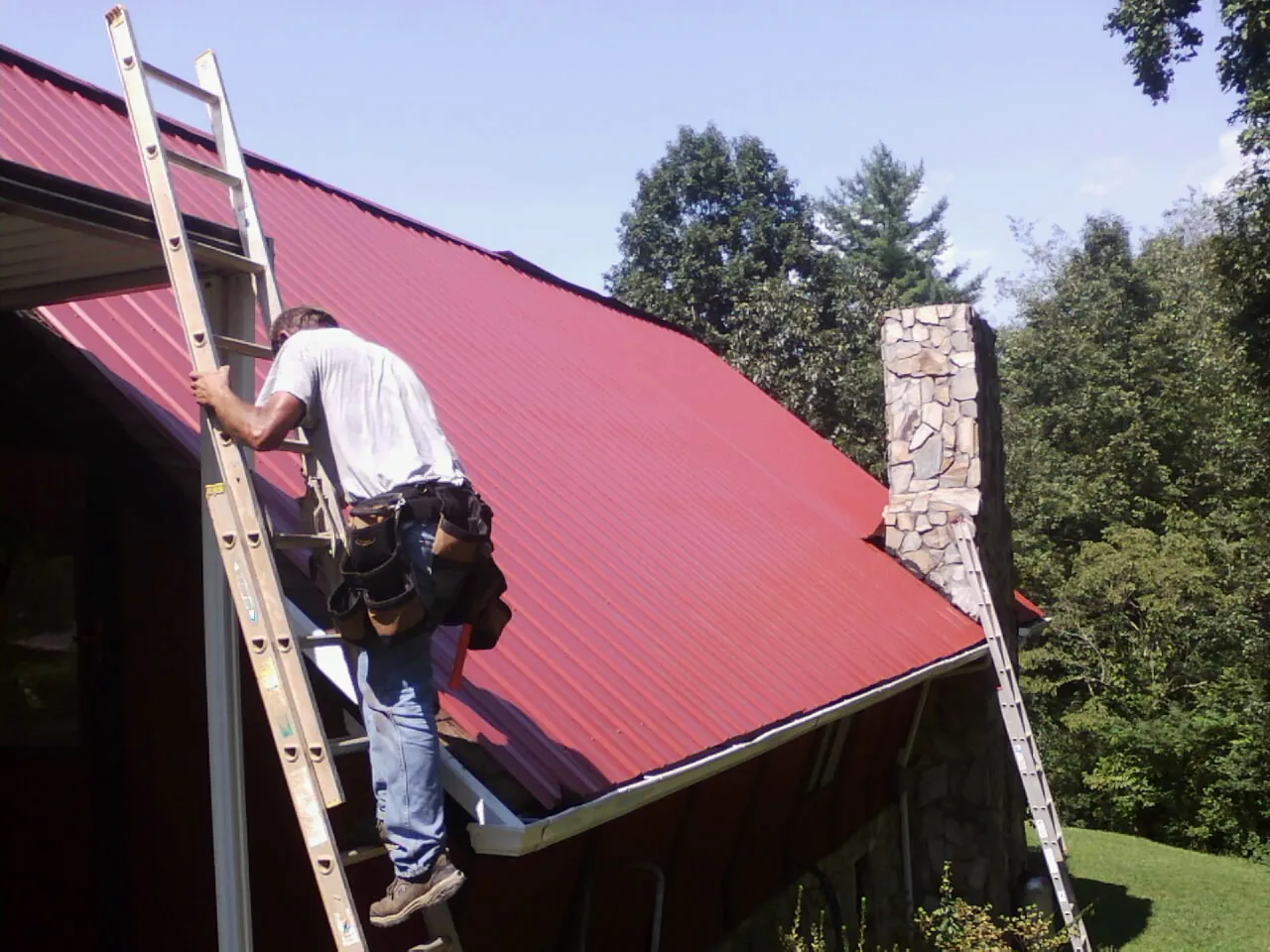 Roofer on a ladder beside a red metal roof and stone chimney — Soffit & Fascia Repair by Roofing Brothers in Water Mill