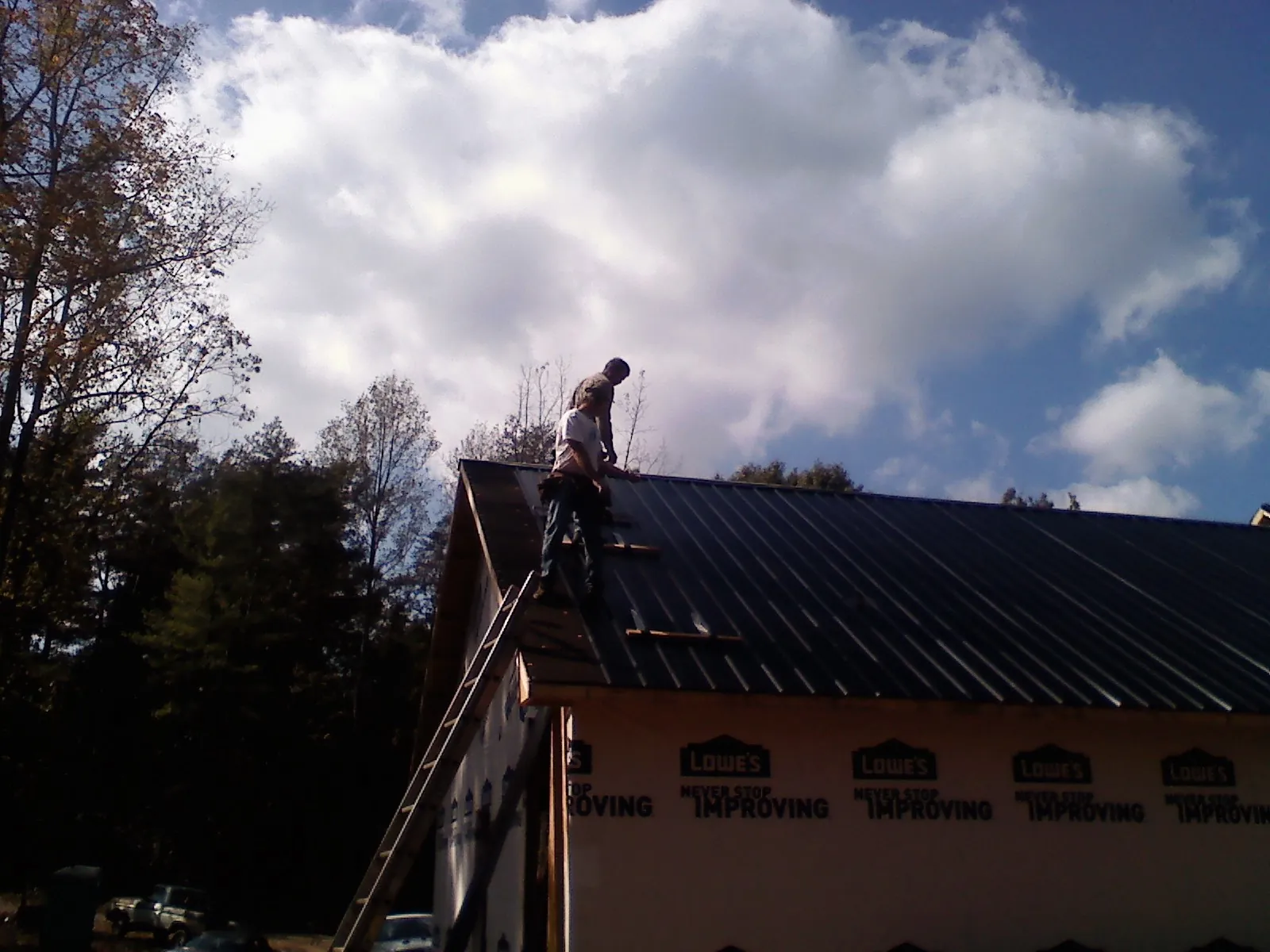 Roofer fastening dark metal panels high against a cloudy sky, from a Roof Inspection job in Water Mill